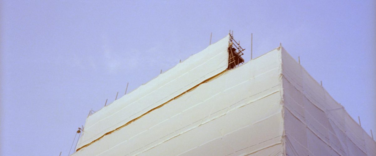 A building under renovation wrapped in protective material with visible scaffolding against a clear sky.